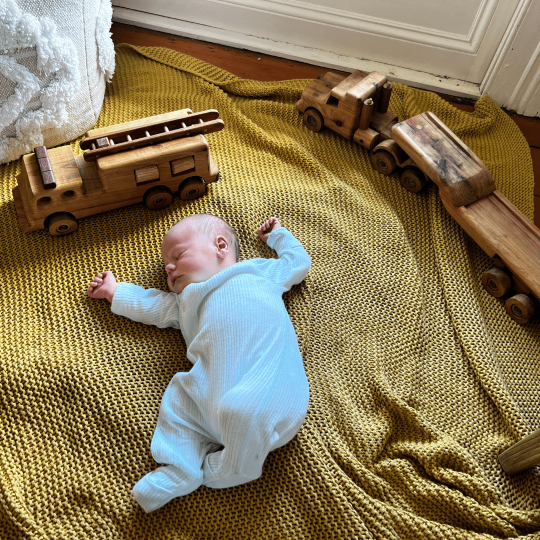 Newborn Baby lying peacefully on mustard knit rug, asleep. Surrounded By Ballard Bear Heirloom Vehicles.