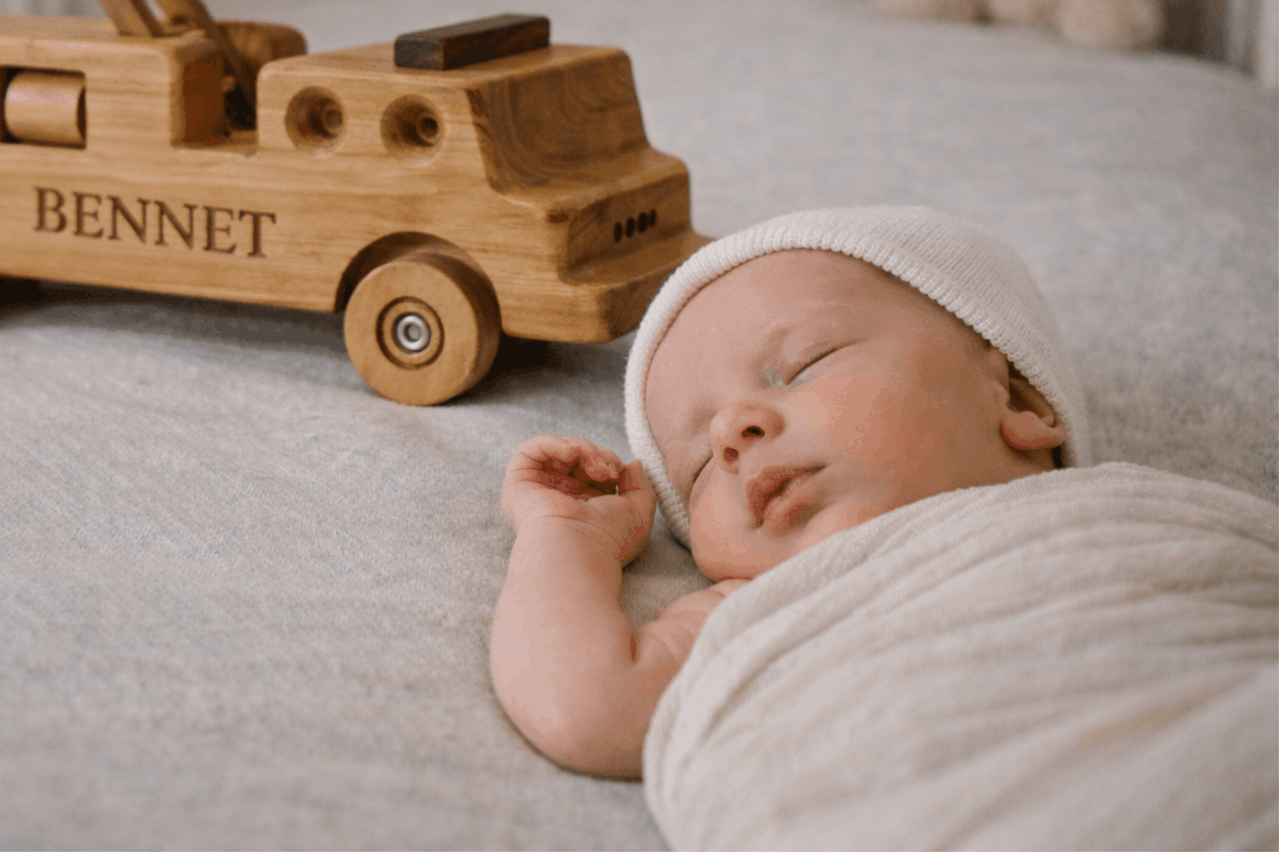 Handcrafted personalised wooden firetruck engraved with the name “Bennett” placed behind a sleeping newborn in a soft neutral nursery, heirloom keepsake gift.