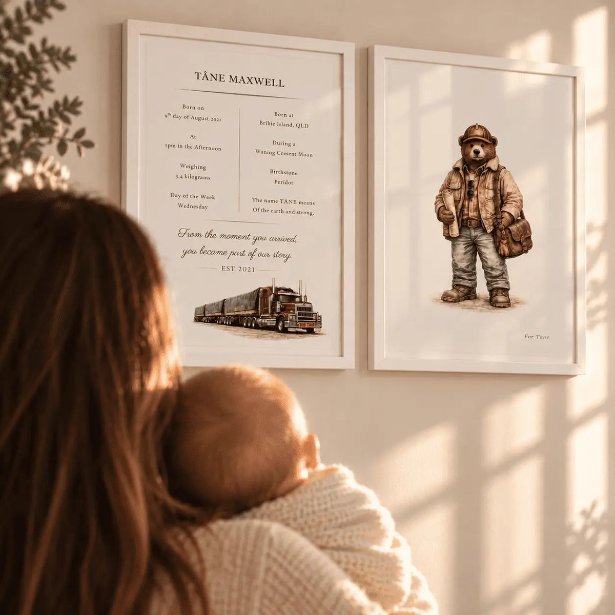 Two framed wall art prints hanging on a sunlit wall, with a woman in a cream knit sweater partially visible in the foreground. The left print reads TÅNE MAXWELL and includes a small road  train illustration; the right print shows a teddy bear in a hat and jacket.