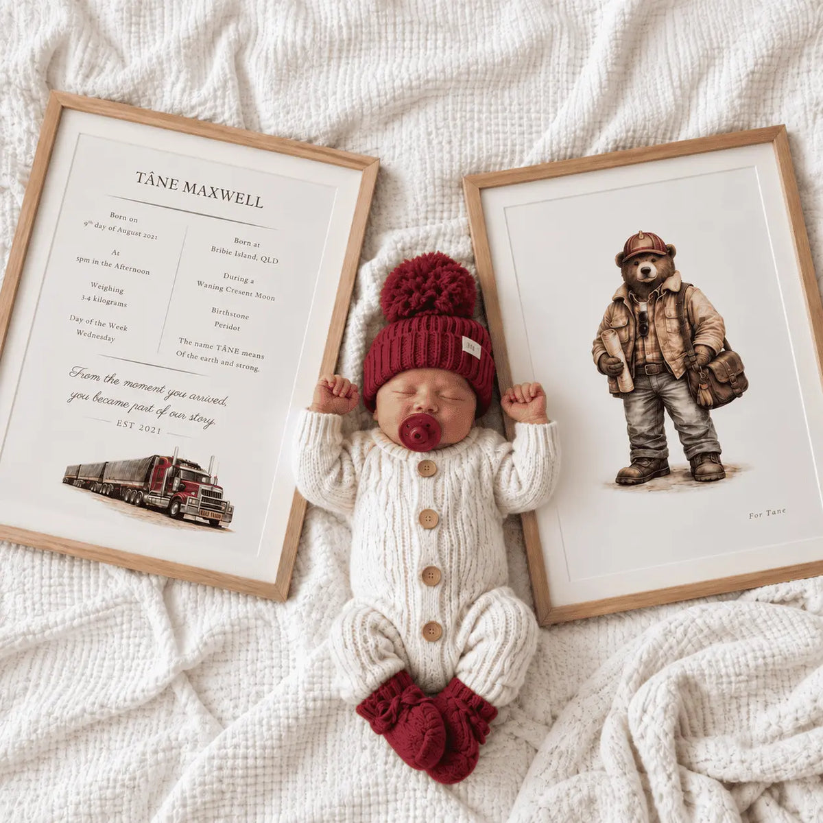 A newborn baby lies on a textured cream blanket between two framed personalized prints. The baby wears a cream knit romper, a dark red knit hat with a pom-pom, and dark red booties. Personalised road train themed birth print with birth details. 