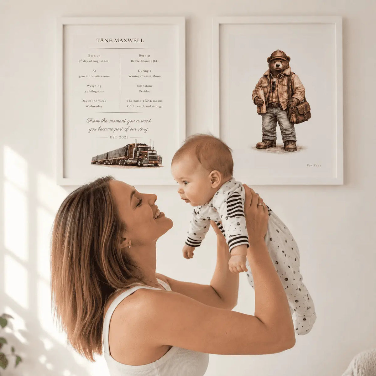 A woman holds a baby in a bright indoor setting, with two framed art prints hanging on the wall behind them. The left print features a road train, personalised birth details and the text 'TÄNE MAXWELL' with a quote, and the right print shows a teddy bear illustration.
