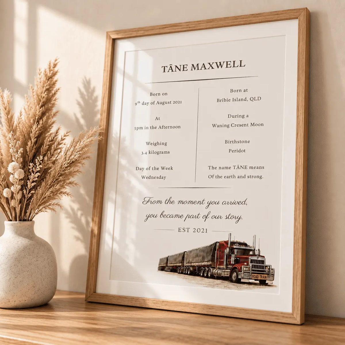 A framed personalized print leaning on a wooden surface beside a vase of dried grass. The print has a light wood frame and a white background with black text reading 'TÄNÄE MAXWELL' and birth details, plus a small illustration of a red semi-truck at the bottom.