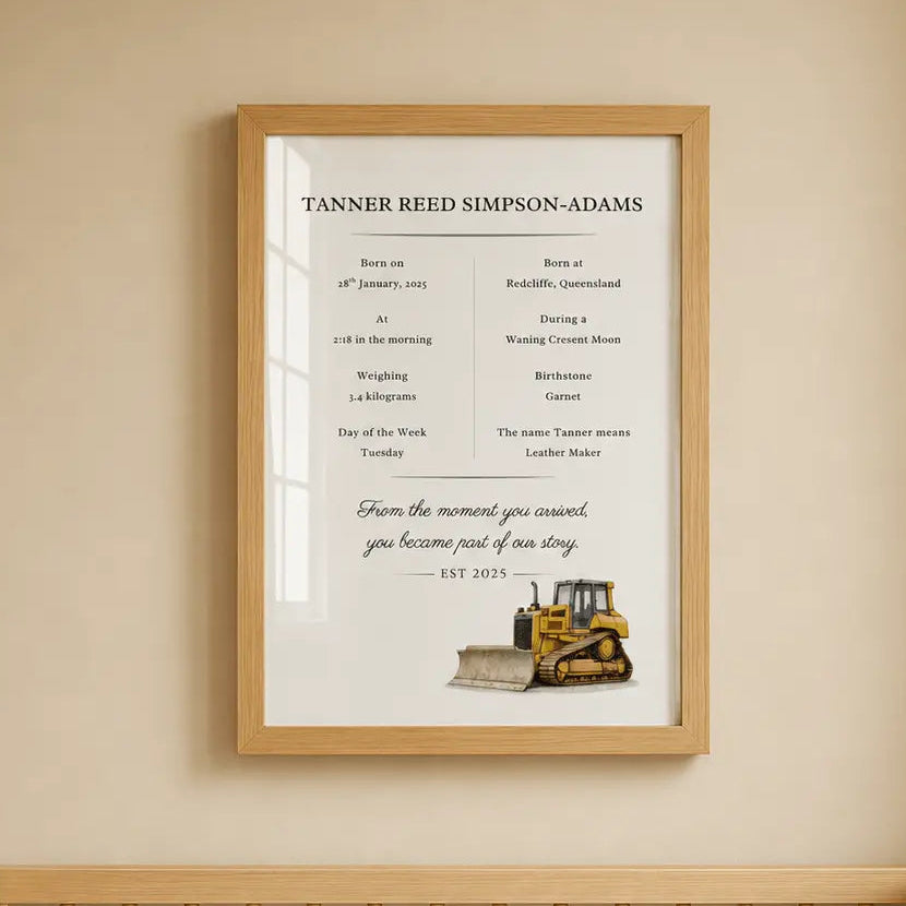 A framed personalized baby nursery print hanging on a beige wall above a wooden crib. The print features a white background with black text detailing a baby's birth and milestones, along with a small illustration of a yellow bulldozer  construction vehicle.
