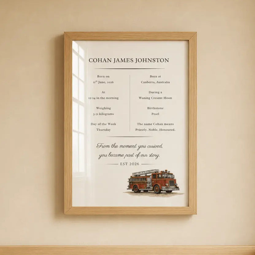 A framed baby birth announcement print hangs on a beige wall above a light wood crib. The print features the baby's birth details in elegant typography and a small illustration of a red fire truck engine at the bottom.